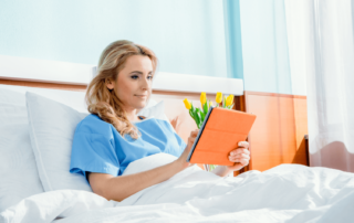 A woman sitting in a hostpital bed after DIEP flap breast reconstruction, reading a book and wearing a blue hospital gown