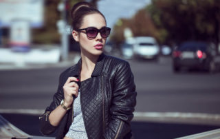 Young fashionable woman standing outside, wearing a black jacket, a grey blouse, dark sunglasses, and with a defined jawline.