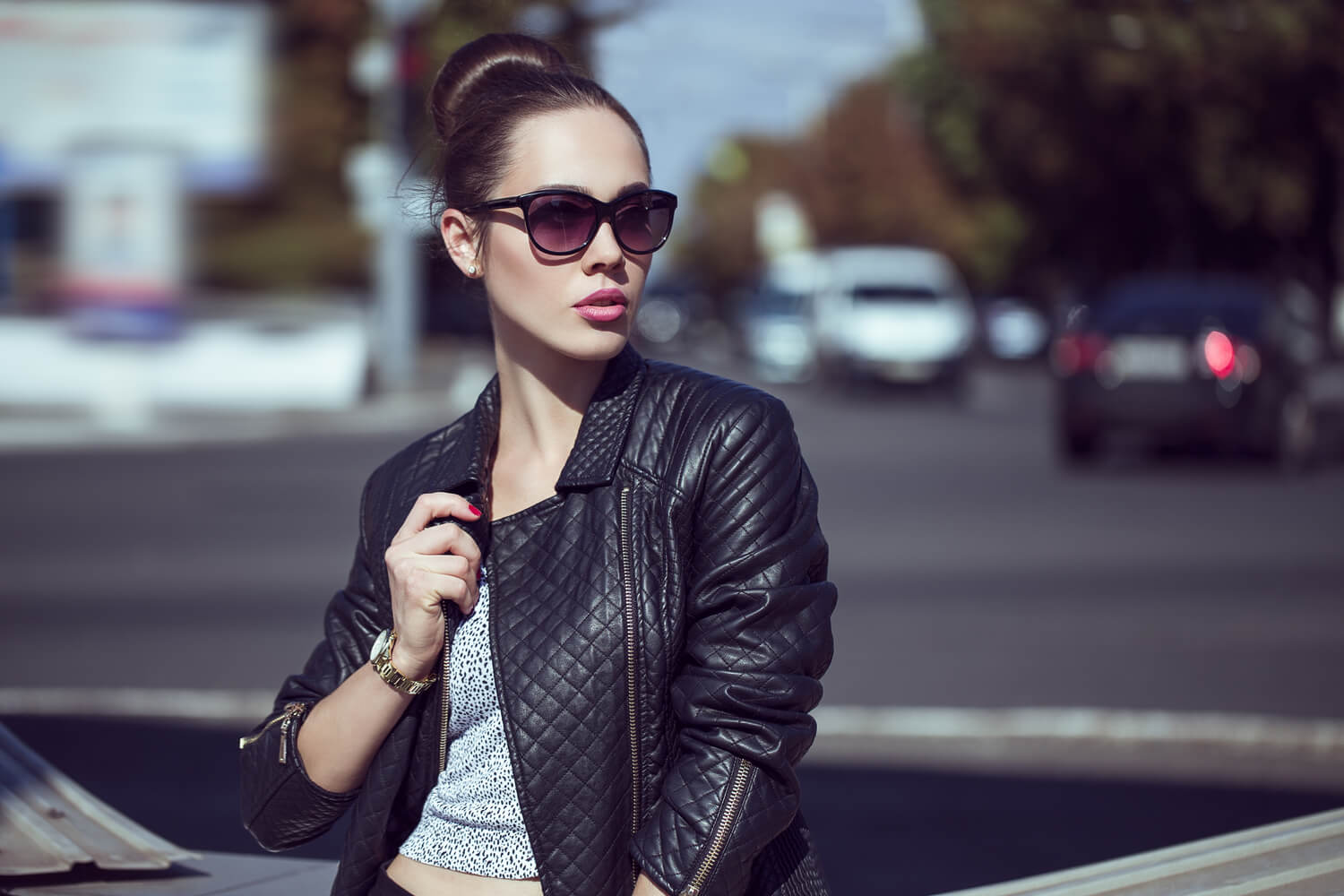 Young fashionable woman standing outside, wearing a black jacket, a grey blouse, dark sunglasses, and with a defined jawline.